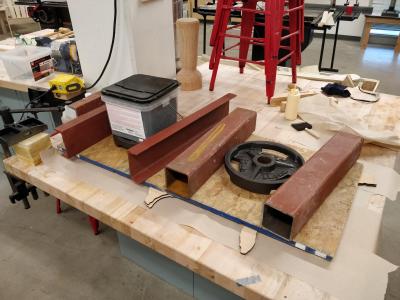 Various pieces of steel and weights stacked on a sheet of chipboard, which is in turned stacked on top of the map.