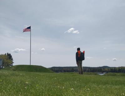 A person standing in a field wearing a backpack carrying four orange gate bundles.